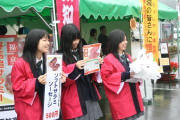 赤い法被を着て、商品のチラシを持った3人の女子高校生がテントの前に並んで、笑顔で販売している写真