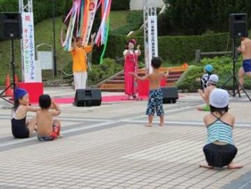 公園内のアスファルトに簡易的な舞台が設けら、歌の宗平お兄さんとみきお姉さんが手拍子をながら歌を歌っており、水着姿の子ども達が集まって、手拍子しながら楽しく参加している様子の写真