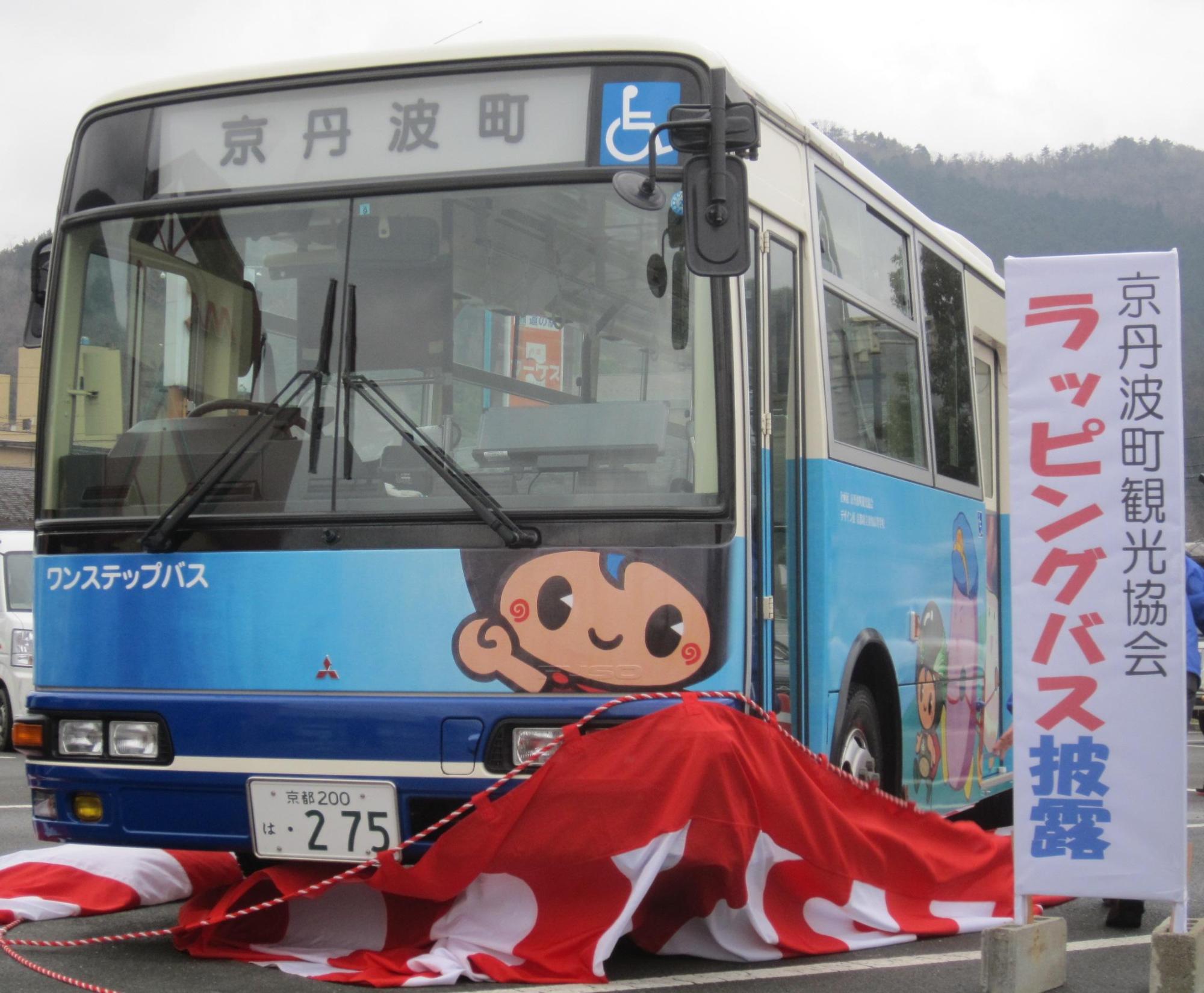 「京丹波町観光協会 ラッピングバス披露」と書かれた看板の横に、紅白の幕が取られ、バスのフロントガラスの下に「味夢くん」のイラストが描かれているラッピングバスの写真