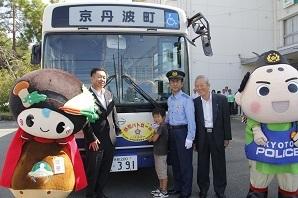バスの正面のフロントガラスの下に「防犯パトロール」と書かれたステッカーが貼られ、運転手さん、町長、男の子、関係者の男性、京丹波町食のキャラクター味夢くん、シンボルマスコットポリスまろんが並んで写っている写真
