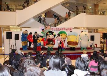 イオンモールの店内に設けられた舞台に、ご当地キャラクターが並んでおり、味夢くんが女性の司会者にインタビューされており、沢山のお客さんが集まっている様子を後方から写した写真