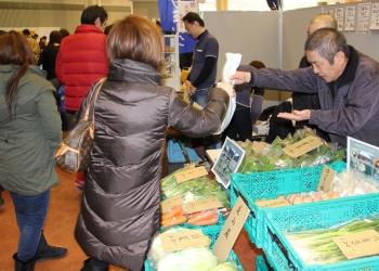 色々な種類の野菜がコンテナに分けて陳列されており、女性のお客さんが、店主から買った野菜が入った袋を受け取っている写真