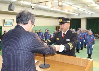 消防の制服と制帽を被った梅原団長が演台の前で紺色のスーツを着た関係者の男性から目録を受け取っており、その様子を後方にいる消防団員の方々が直立で見守っている写真