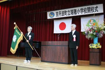 「京丹波町立瑞穂小学校開校式」と書かれた横断幕、日の丸の国旗、京丹波町章が舞台上に掲げられ、胸に胸章を付けた男性2名が舞台に立っており、左側の男性が緑色の学校旗を持っている開校式の写真