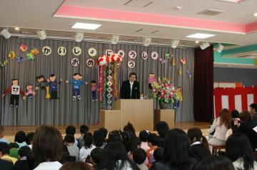 舞台後方の幕に「かいしょ にゅうしょ おめでとう」の文字と友達が手をつないでいる壁面装飾がされ、演台の前に立って話をしている寺尾町長と開所式に参加している園児や保護者の方々が座って話を聞いている「みずほ保育所」開所式の写真