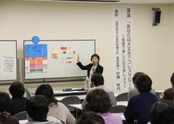 資料を掲示したホワイドボード、マイクを持って話をしている管理栄養士の中澤幸代さん、参加者が長机の席に着いて講座を受けている様子を後方から写した写真