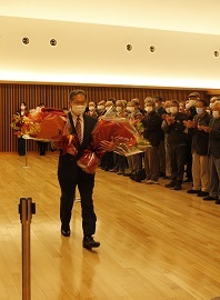 町民や議員、職員の方々が拍手をしており、畠中町長が両手に大きな花束を持って歩いている写真