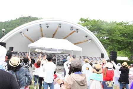 野外の半円型の屋根のあるステージに向かって沢山の観客が両手を上に挙げて手拍子をするなどしてロックフェスティバルを楽しんでいる様子を後方から写した写真