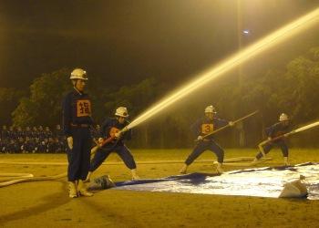 ライトが照らされている夜に、消防団員の方が足を大きく前後に広げ、腰を少し落とし、両手で消防ホースを持ち構えて放水し、勢いよく水が噴き出している訓練を披露している写真