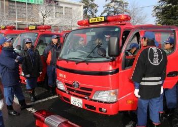 赤い消防車両が数台並んで駐車されており、一台の消防車両の運転席の両側の扉を開けて、消防団員が運転席の中の様子を見て、確認している写真