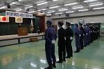 舞台には日の丸の国旗、京丹波町章、横断幕が掲げられ、演台の前に立って話をしいている男性、消防団員の方々が手を後ろに組んで直立し、横一列に並んでいる町消防団入退団および役員任命式の写真