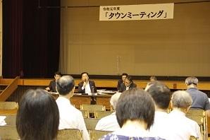 「タウンミーティング」と書かれた横断幕、前方に設置され長机、町長と職員の方が座り、中央に座っている町長がマイクを持って話をしており、たくさんの参加者が席に着き、話を聞いている写真