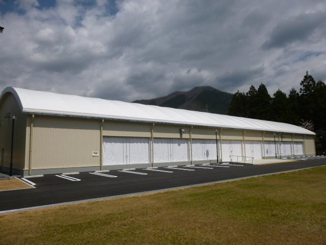 手前に芝生の広場や駐車場がある横長の白いアーチ型の屋根付き屋内多目的広場の外観写真